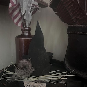 Op de afbeelding: Een rustieke display met een kleine Amerikaanse vlag, veren en een silhouet van een heksenhoed. Een bruine fles bevat de vlag en veren. Een klein bordje leest "Salem 1691". De scène roept een vintage, historisch thema op.