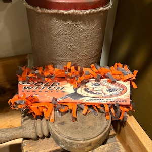 Op de afbeelding: Een vintage Old Witch sterretjesdoos met een afbeelding van een heksengezicht, omringd door oranje en zwarte papieren franjes. De doos staat voor een cilindrische container met een rood deksel en een houten voet met een handvat.