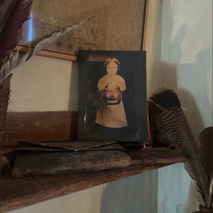 Op de afbeelding: Een vintage foto van een jong kind dat een gesneden houten pompoen vasthoudt. De foto is ingelijst in een donkerhouten lijst en wordt tentoongesteld op een houten plank met andere antieke voorwerpen.