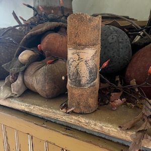 Puede incluir: Una vela cilíndrica marrón texturizada con una ilustración vintage de una calabaza en la parte delantera. La vela está rodeada de calabazas de tela y otra decoración de otoño.