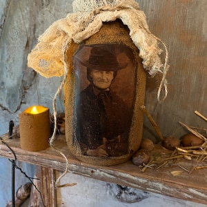 Op de afbeelding: Een decoratieve pot met een vintage portret van een persoon met een hoed en een kat. De pot is bedekt met een textuurrijk materiaal en afgewerkt met een beige stof. Een kleine kaars staat links.