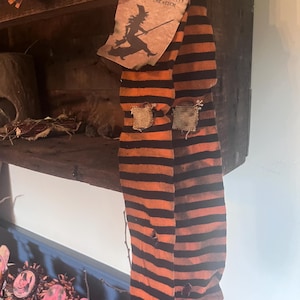 Op de afbeelding: Een paar oranje en zwart gestreepte sokken met een versleten uiterlijk, kleine gaatjes en lapjes. Een rechthoekig papieren label is aan de bovenkant bevestigd en toont de tekst HALLOWEEN SEASON OF THE WITCH en het silhouet van een heks.