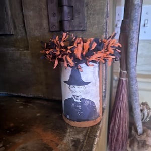Könnte beinhalten: Zylindrische Halloween-Dekoration mit einem Schwarz-Weiß-Bild eines Mannes mit Hut. Die Oberseite des Zylinders ist mit orangefarbenem und schwarzem Fransenstoff verziert. Die Dekoration steht auf einer Holzoberfläche.