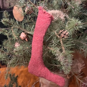 May include: A handmade red stocking ornament hanging on a small pine tree. The stocking is made of a soft, textured fabric and has a rustic, vintage look.