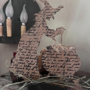 May include: A wooden witch silhouette with a cauldron, featuring script-like text. The witch is brown with a black base, and the cauldron has straw and a handle. The background includes a wall sconce with three light bulbs.
