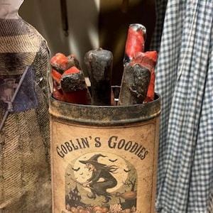 May include: A cylindrical metal container labeled Goblin's Goodies Old Fashioned Halloween Candy Est. 1898, featuring a graphic of a witch on a broomstick. The container is filled with several dark, textured, red and black cylindrical objects.