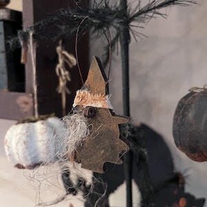 May include: A rustic Halloween tree ornament shaped like a witch, crafted from dark brown material. The witch has a pointed hat, a white beard, and a tan fabric band. Other Halloween-themed ornaments are also visible.