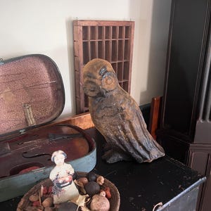 Op de afbeelding: Een verweerde, bruine uil standbeeld staat op een zwart oppervlak, naast een vintage vioolkist en een geweven mand gevuld met decoratieve items. Een houten vitrinekast hangt op de achtergrond, wat bijdraagt aan de antieke esthetiek.