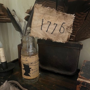 Puede incluir: Una exhibición de estilo vintage con una botella de vidrio transparente, una bandera de arpillera con el número "1776" y una bolsa de cuero oscuro. La escena incluye plumas y un pequeño libro, evocando un tema histórico.