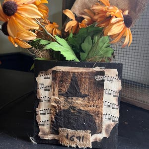 Op de afbeelding: Een zwarte houten doos met een collage van vintage papier en stof. De collage toont een silhouet van een heks met een hoed en het jaar 1691. De doos is versierd met een boeket oranje en gele bloemen.