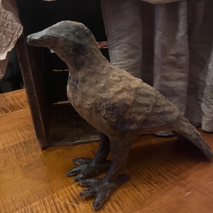 Op de afbeelding: Een vogelbeeldje met een getextureerde, verweerde bruine en zwarte afwerking. Het beeldje heeft een spitse snavel, gedetailleerde vleugels en donkere, opvallende poten. Het oppervlak heeft een matte, verouderde uitstraling met kleurvariaties.