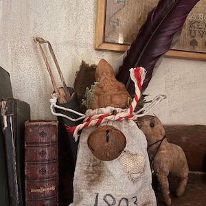 May include: A small, rustic, brown gnome figurine is nestled in a burlap sack with a red and white striped ribbon. The sack has a rusty bell attached and the year "1803" is written on the front.