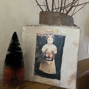 Op de afbeelding: Een vintage Halloween-kunstwerk op canvas met een verweerde witte achtergrond. Het canvas toont een zwart-witfoto van een kind dat een jack-o'-lantern vasthoudt met de woorden "Happy Halloween" erop.