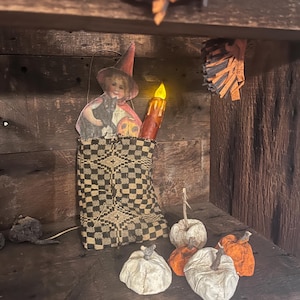 Op de afbeelding: Een Halloween-display met een brandende kaars, een heks en zwarte kat uitsparing en kleine pompoenen. De pompoenen zijn wit en oranje. De display staat op een houten plank.