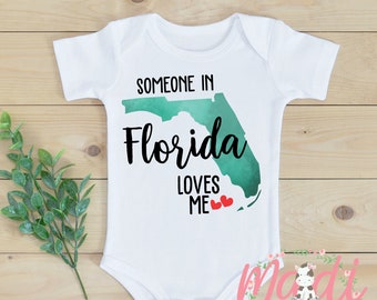 May include: White baby onesie with a green watercolor outline of the state of Florida and the text "Someone in Florida loves me" with two red hearts.