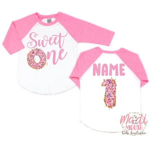 May include: Pink and white baseball style shirt with the text "Sweet One" and a pink donut with sprinkles on the front. The back of the shirt has the text "NAME" and a pink donut shaped like the number one with sprinkles.