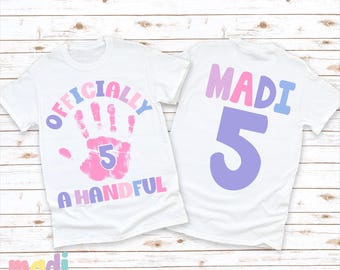 May include: Two white t-shirts with a pink handprint and the text "Officially a Handful" on one shirt and the text "Madi 5" on the other shirt.