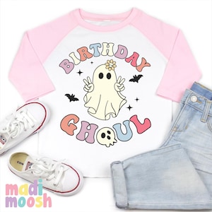 May include: A white and pink baseball tee shirt with a ghost wearing a flower on its head and the text "Birthday Ghoul" in rainbow colours.