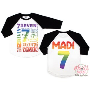 Peut inclure: Un t-shirt de baseball blanc et noir avec du texte de couleur arc-en-ciel et un grand num&eacute;ro 7 arc-en-ciel au dos. Le devant du t-shirt dit "7SEVEN" plusieurs fois dans diff&eacute;rentes polices et tailles. Le dos du t-shirt dit "MADI" avec un num&eacute;ro 7 arc-en-ciel en dessous. Le t-shirt est pour un enfant.