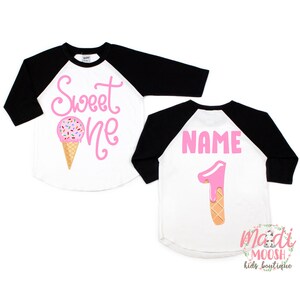 May include: A white and black baseball tee shirt with pink text that says "Sweet One" and an ice cream cone graphic. The back of the shirt has a pink number "1" with pink icing and a waffle cone, and the text "NAME" in pink.