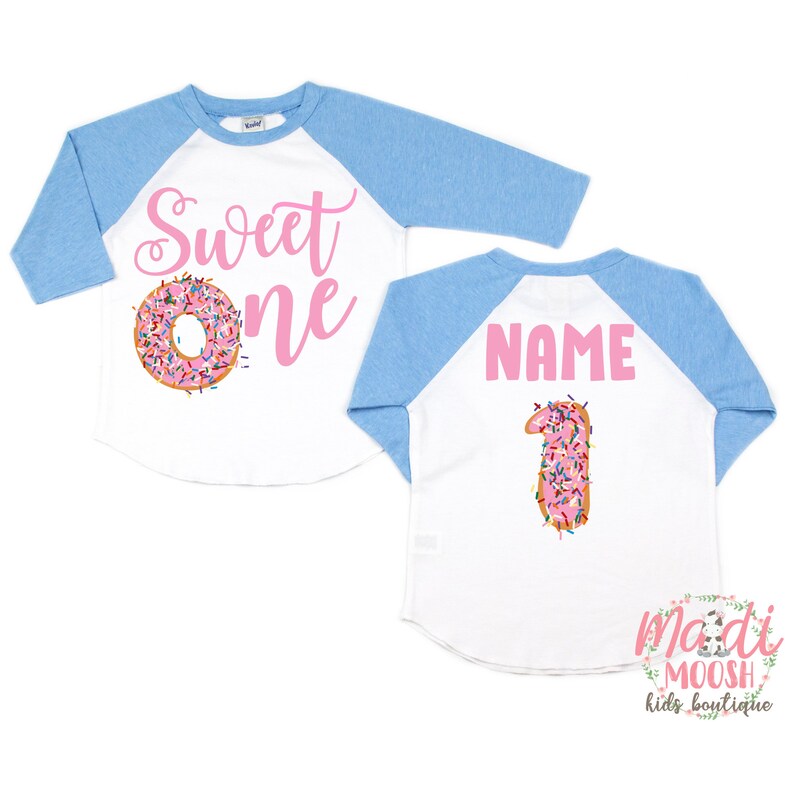 May include: A white and blue baseball style shirt with the words "Sweet One" in pink script and a pink frosted donut with sprinkles on the front. The back of the shirt has the word "NAME" in pink script and a pink frosted donut with sprinkles shaped like the number one.