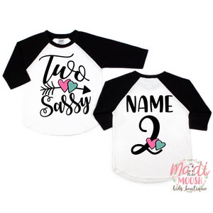 May include: White and black raglan style shirt with the text "Two Sassy" and an arrow with pink and teal hearts. The back of the shirt has the text "Name" and the number "2" with pink and teal hearts. The shirt is from Madi Moosh Kids Boutique.