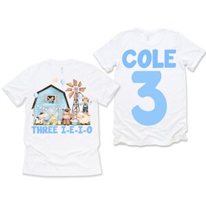 May include: White t-shirts with a farm theme. One shirt features a blue barn, windmill, and farm animals with the text "THREE I-E-I-O". The other shirt has the name "COLE" and the number "3" in blue.