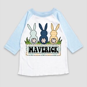 May include: A white and light blue baseball tee shirt with a design of three bunnies with different colored patchwork patterns. The bunnies are sitting on a patch of green grass with a banner that says "MAVERICK" in black letters.