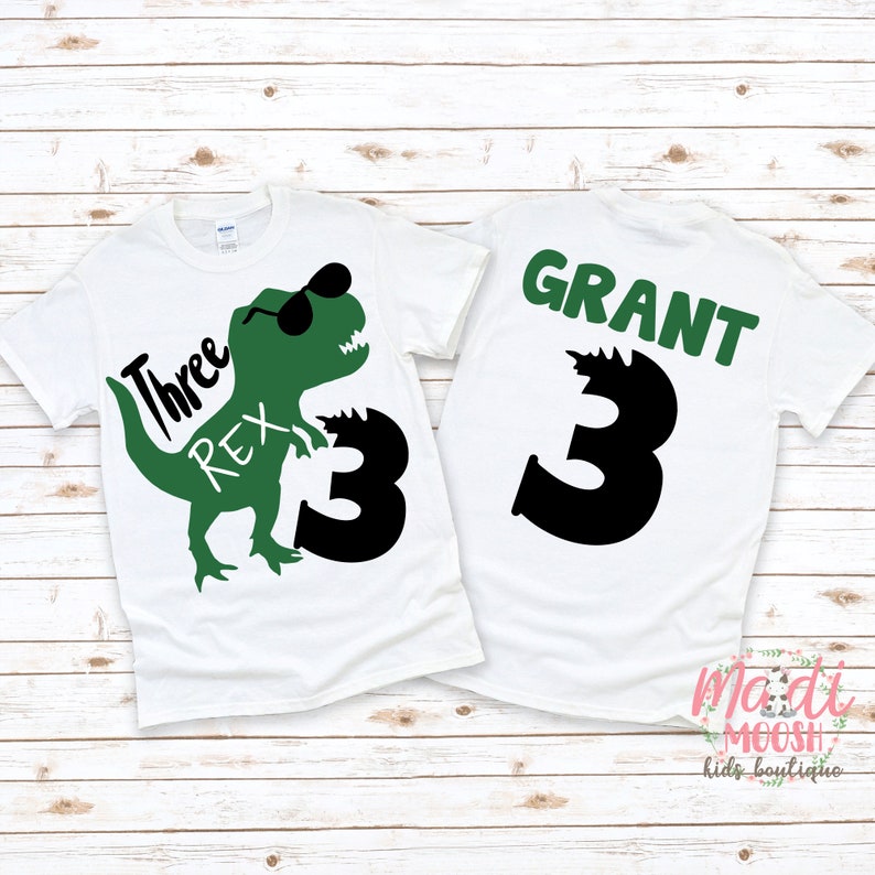 Op de afbeelding: Twee witte T-shirts met een groene dinosaurus afbeelding en de tekst "Three Rex" op het ene shirt en de tekst "GRANT" en het nummer "3" op het andere shirt.