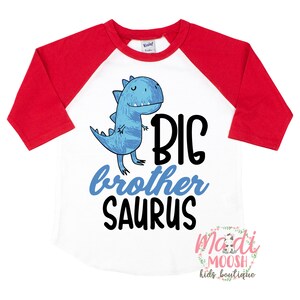 May include: White and red baseball tee shirt with a blue dinosaur graphic and the text "BIG brother SAURUS". The shirt has a white background with red sleeves and a white collar. The text is in black. The dinosaur is blue and has a friendly expression. The text "madi MOOSH kids boutique" is in the bottom right corner.