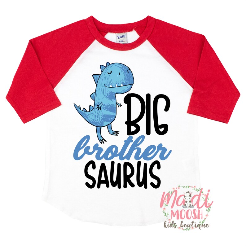 May include: White and red baseball tee shirt with a blue dinosaur graphic and the text "BIG brother SAURUS". The shirt has a white background with red sleeves and a white collar. The text is in black. The dinosaur is blue and has a friendly expression. The text "madi MOOSH kids boutique" is in the bottom right corner.