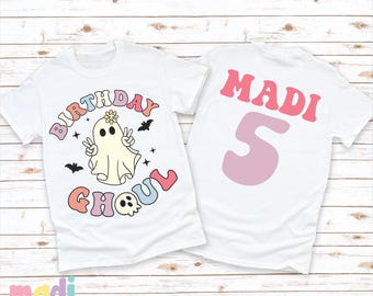May include: White t-shirt with a ghost wearing a flower crown and the text "Birthday Ghoul" in rainbow colors. The back of the shirt has the name "MADI" and the number "5" in pink.