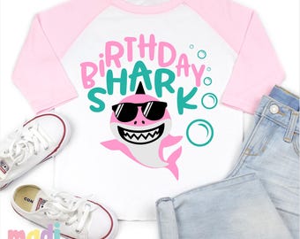 May include: White and pink raglan-style t-shirt with the words "Birthday Shark" and a cartoon shark wearing sunglasses. The shirt is paired with light blue denim shorts and white sneakers.
