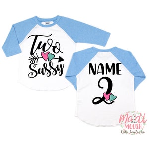 May include: A white and light blue raglan shirt with the text "Two Sassy" and an arrow graphic on the front. The back of the shirt has the word "NAME" above the number "2". Pink and teal hearts are also on the shirt.