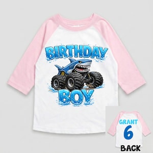 May include: White and pink raglan-style shirt with a blue shark monster truck graphic. The front of the shirt says "BIRTHDAY BOY" in blue lettering. The back of the shirt has the name "GRANT" and the number "6".