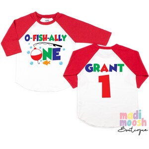 May include: A white and red raglan style shirt with the text "O-Fish-Ally One" and a fishing theme graphic on the front. The back of the shirt has the name "Grant" and the number "1" printed on it.