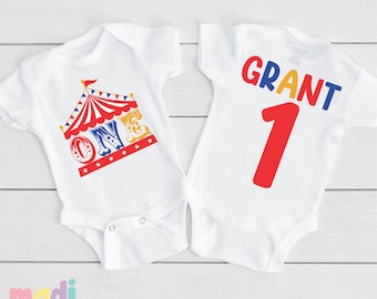 May include: White baby onesie with a red, yellow, and blue circus tent and the word "ONE" in colorful letters on the front. The back of the onesie has the name "GRANT" in yellow, red, and blue letters and the number "1" in red.