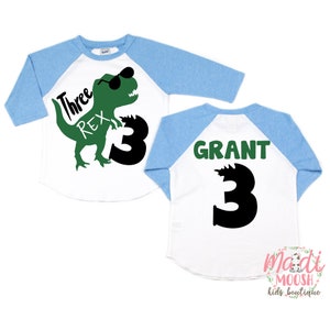 Op de afbeelding: Een wit en blauw baseballshirt met een groene dinosaurus die een zonnebril draagt en de tekst "Three Rex" op de voorkant. De achterkant van het shirt heeft de naam "GRANT" en het nummer "3" in groen.