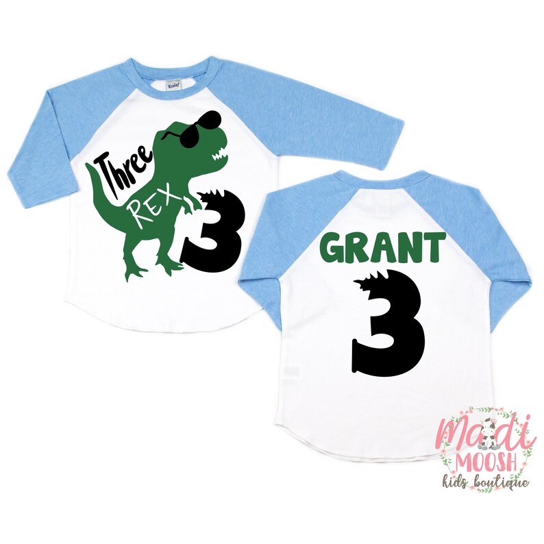 Op de afbeelding: Een wit en blauw baseballshirt met een groene dinosaurus die een zonnebril draagt en de tekst "Three Rex" op de voorkant. De achterkant van het shirt heeft de naam "GRANT" en het nummer "3" in groen.