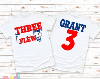 May include: Two white t-shirts with red and blue text. The front shirt says "Three years flew by" with a small airplane graphic. The back shirt says "Grant 3".