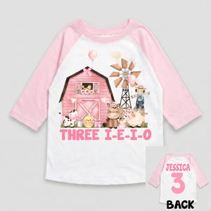 May include: A white and pink raglan-style shirt with a farm-themed graphic. The front features a pink barn, windmill, and cartoon animals. The text "THREE I-E-I-O" is displayed. The back of the shirt has the name "JESSICA", the number "3", and the word "BACK".