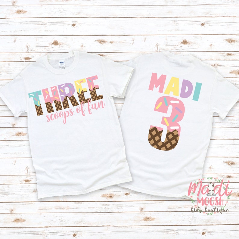 May include: White t-shirt with the text "THREE scoops of fun" in pink, yellow, and blue with a brown waffle cone and sprinkles. The back of the shirt has the name "MADI" in pink, yellow, and blue with the number "3" in pink with a brown waffle cone and sprinkles.