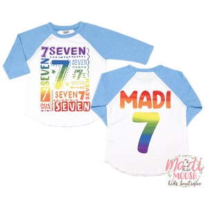 Peut inclure: Un t-shirt de style baseball blanc et bleu avec un num&eacute;ro sept arc-en-ciel et le mot "SEVEN" r&eacute;p&eacute;t&eacute; en diff&eacute;rentes tailles et couleurs sur le devant. Le dos du t-shirt porte le nom "MADI" et un num&eacute;ro sept arc-en-ciel.