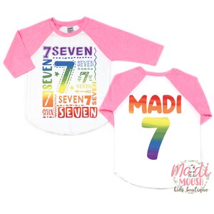 Peut inclure: Un t-shirt de baseball blanc et rose avec un motif arc-en-ciel repr&eacute;sentant le chiffre 7 et le mot "SEVEN" r&eacute;p&eacute;t&eacute; plusieurs fois. Le dos du t-shirt porte le nom "MADI" et le chiffre 7 en couleurs arc-en-ciel.