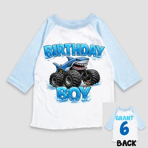 May include: A white and light blue raglan-sleeve shirt with a shark-themed monster truck graphic. The graphic includes the words "BIRTHDAY BOY". The back of the shirt has the name "GRANT", the number "6", and the word "BACK".
