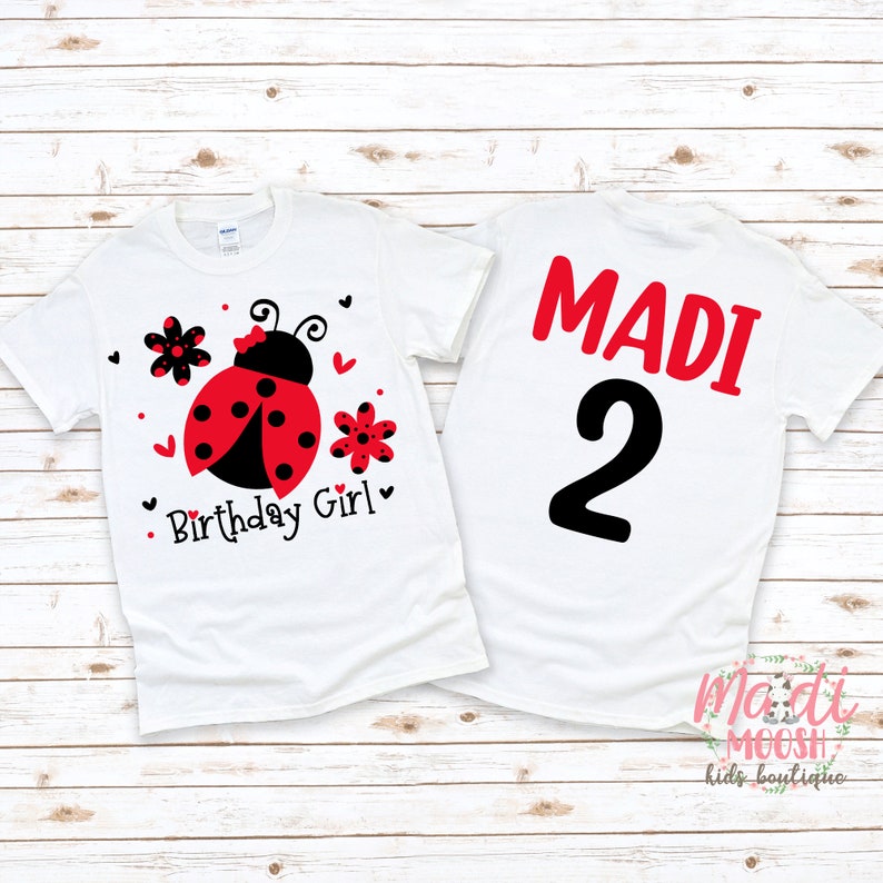 May include: White t-shirt with a red ladybug, flowers, and the text "Birthday Girl". The back of the shirt has the name "MADI" and the number "2" in black.