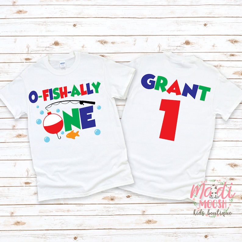 May include: Two white t-shirts with colorful graphics. One shirt says "O-FISH-ALLY ONE" with a fishing bobber, fish, and bubbles. The other shirt has the name "GRANT" and the number "1" on the back. The shirts are on a light wood background.