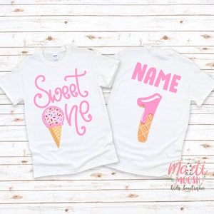 May include: White t-shirt with pink text that says "Sweet One" and a pink ice cream cone with sprinkles. The back of the shirt has a pink number "1" shaped like an ice cream cone with the text "NAME" above it.