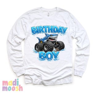 May include: White long-sleeve shirt with a graphic of a blue shark monster truck. The text "BIRTHDAY BOY" is in blue, with water splash accents. The shirt has a crew neck and long sleeves.