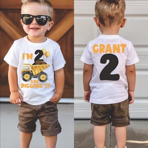 May include: White t-shirt with a yellow dump truck and the text "I'm 2 and digging it". The back of the shirt has the name "GRANT" and the number "2" in black.